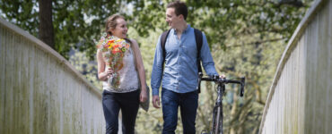 James & Ali walking across a bridge holding flowers & wheeling a bike.