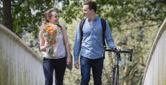 James & Ali walking across a bridge holding flowers & wheeling a bike.