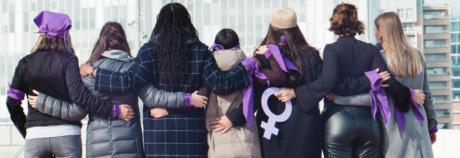 Group of women, backs to the camera, arms linked, wearing purple for International Women's Day
