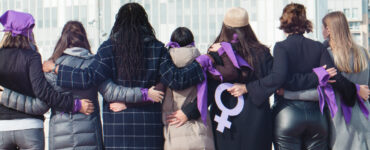 Group of women, backs to the camera, arms linked, wearing purple for International Women's Day