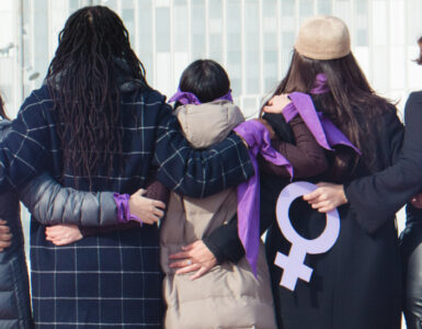 Group of women, backs to the camera, arms linked, wearing purple for International Women's Day