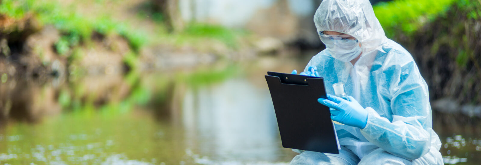 Female scientist in a white protective suit crouched in a river making notes