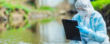 Female scientist in a white protective suit crouched in a river making notes