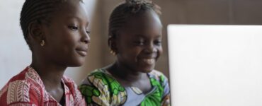 Two girls in Ghana sat in front of a laptop smiling
