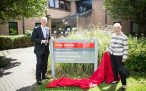 Polly standing next to a sign for the Polly Vacher building with the then Vice-Chancellor David Bell