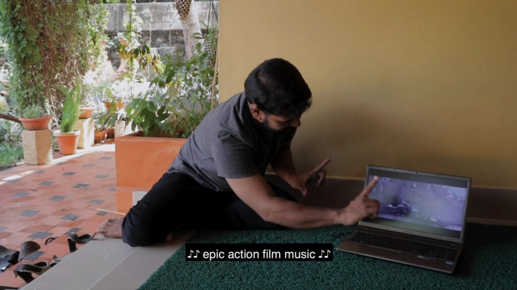  A man wearing a grey T-shirt and black pants against a yellow wall. He is sitting on the floor next to his laptop, with his hands making a film frame as he discusses a shot from a film playing on his laptop. The caption reads [Epic action film music].