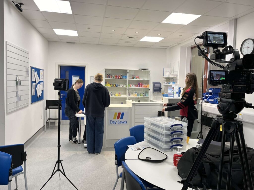 Pharmacy-style room with people working at a counter labeled 'Day Lewis'; camera setup for filming.