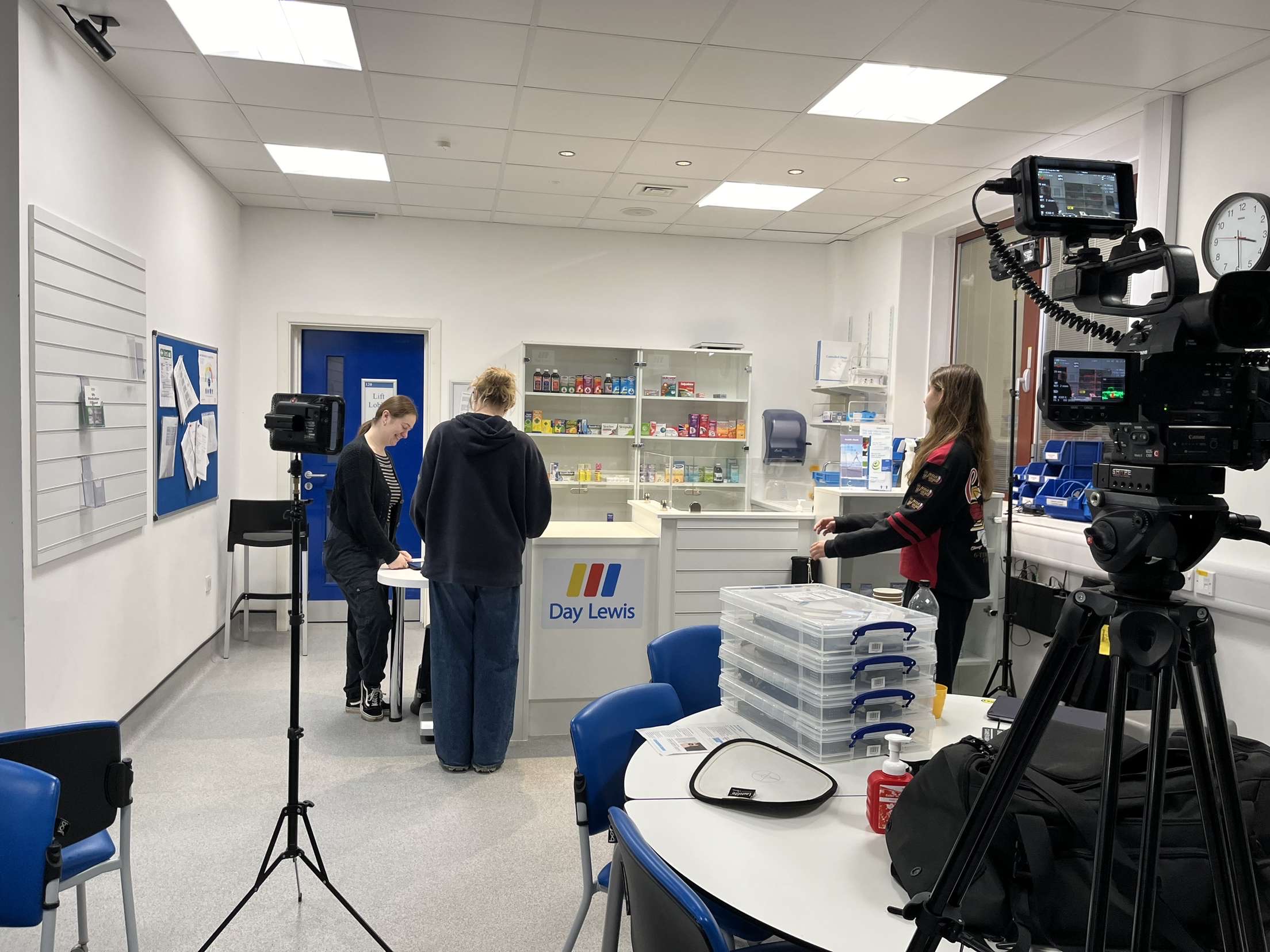 Pharmacy-style room with people working at a counter labeled 'Day Lewis'; camera setup for filming.