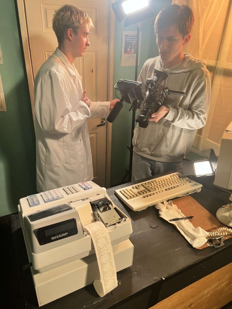 Two people filming near a table with an old cash register, keyboard, and papers under studio lights.