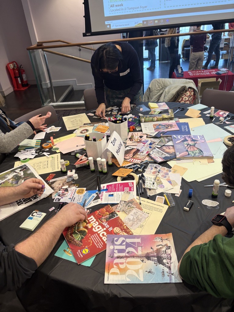 People working on a zine-making project at a round table covered with magazines, paper, glue sticks, and craft supplies.