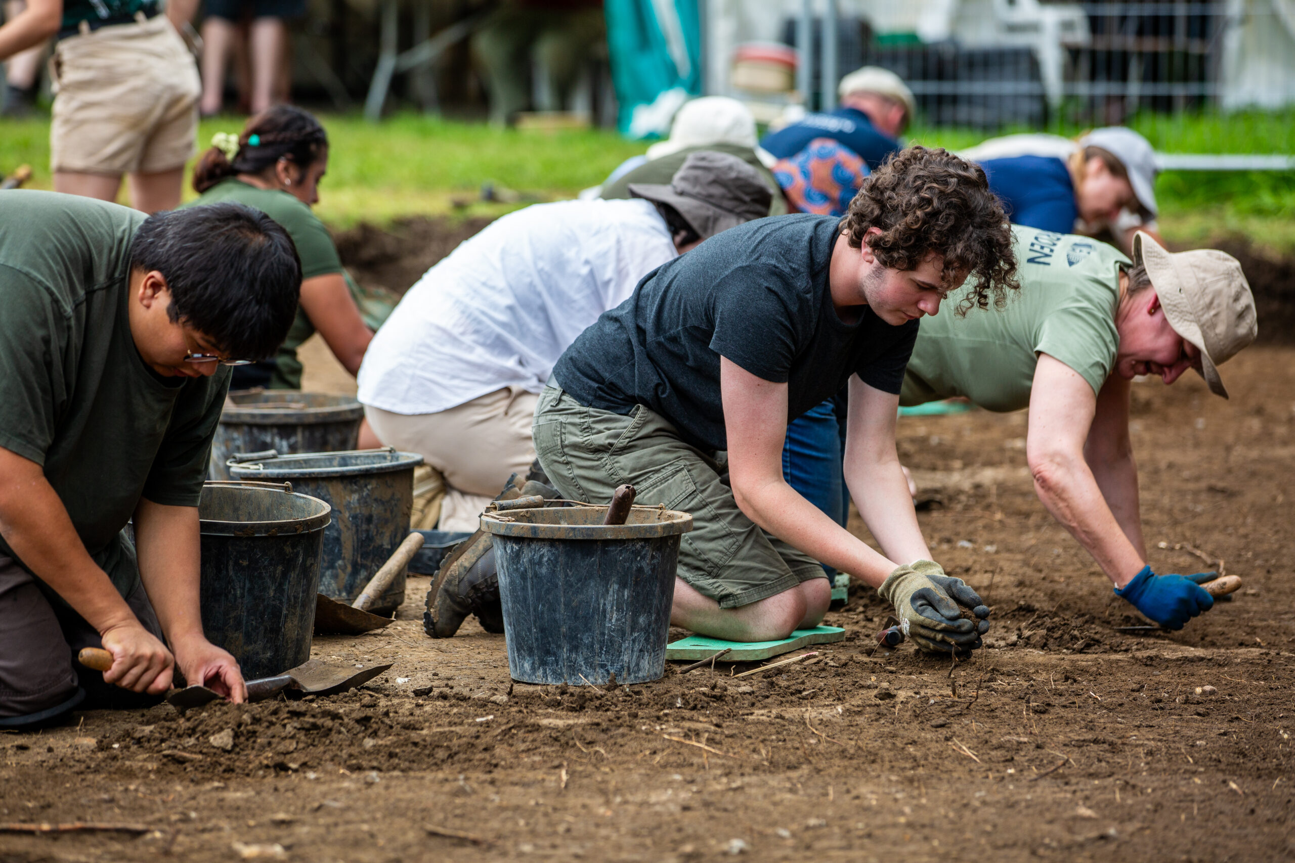 A group of people are kneeling and crouching in a row outdoors, engaged in an archaeological dig. They are working close to the ground, using small hand tools to scrape and uncover soil. Several black plastic buckets are placed in front of them for collecting material. The ground is a cleared dirt area, and the background shows greenery, equipment, and partially visible structures indicating an active dig site.