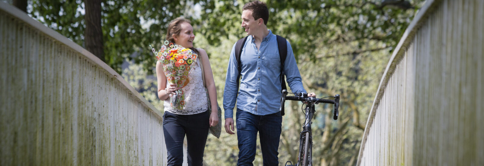 James & Ali walking across a bridge holding flowers & wheeling a bike.