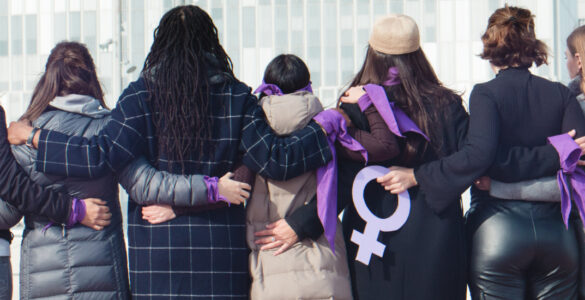 Group of women, backs to the camera, arms linked, wearing purple for International Women's Day